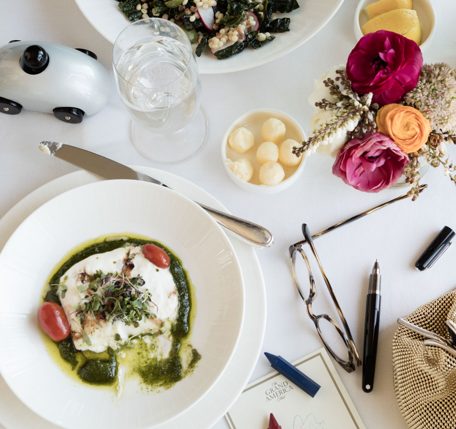 Grand America table setting with cheese and salad in a white bowl and handbag, toy car, and Grand America Hotel notepad.