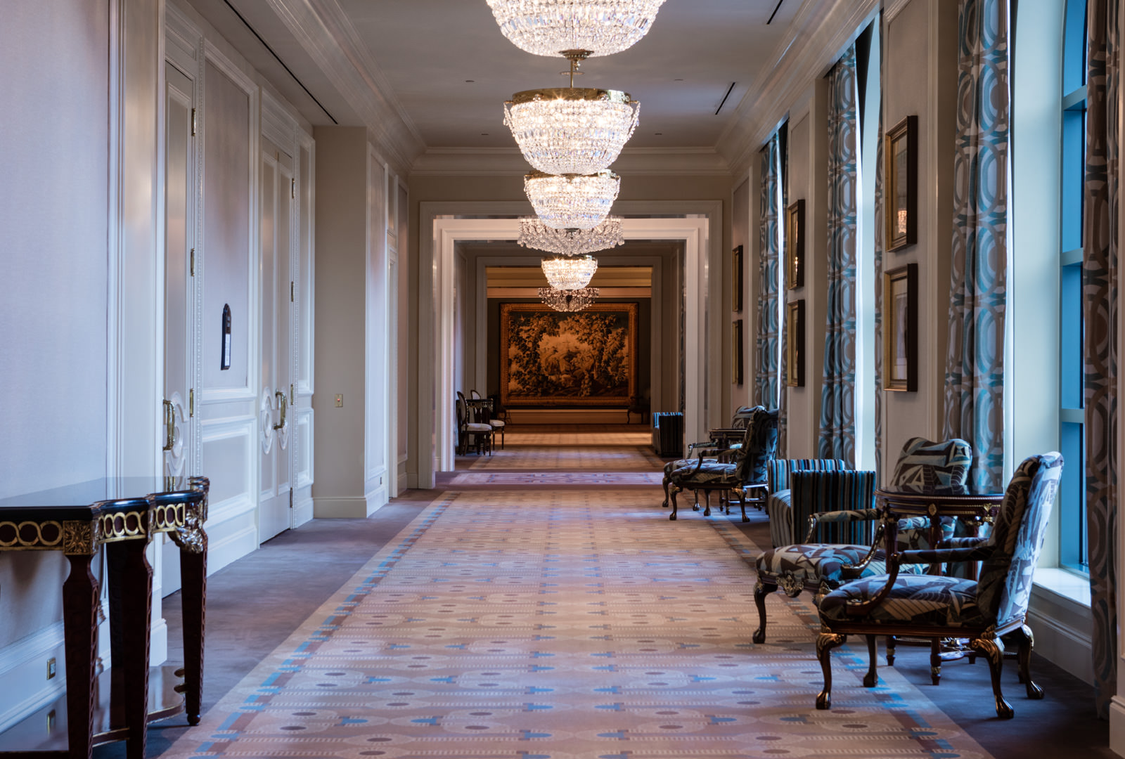 Long hotel hallway with chandeliers, blue curtains, and a tapestry at the end