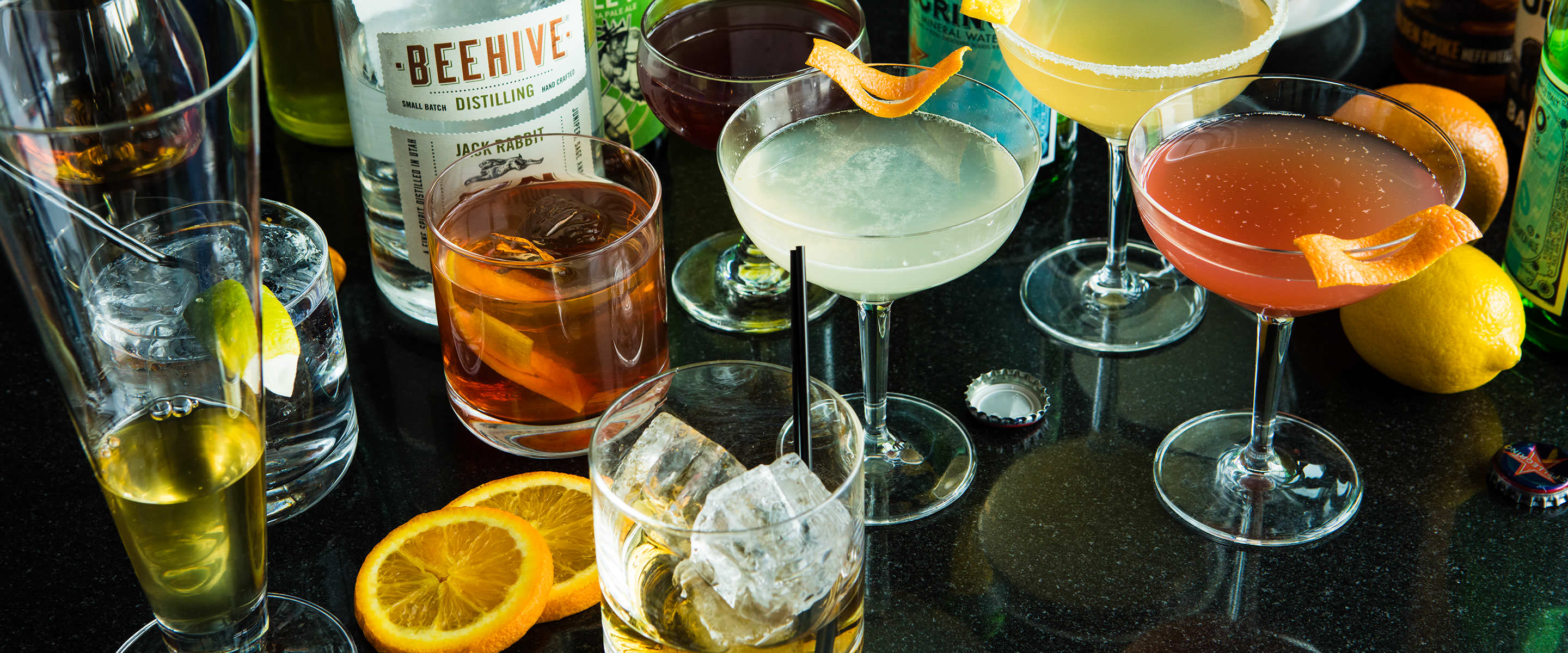 Cocktail drink display with liquor bottles, wine glasses, tumbler glasses and orange garnish.