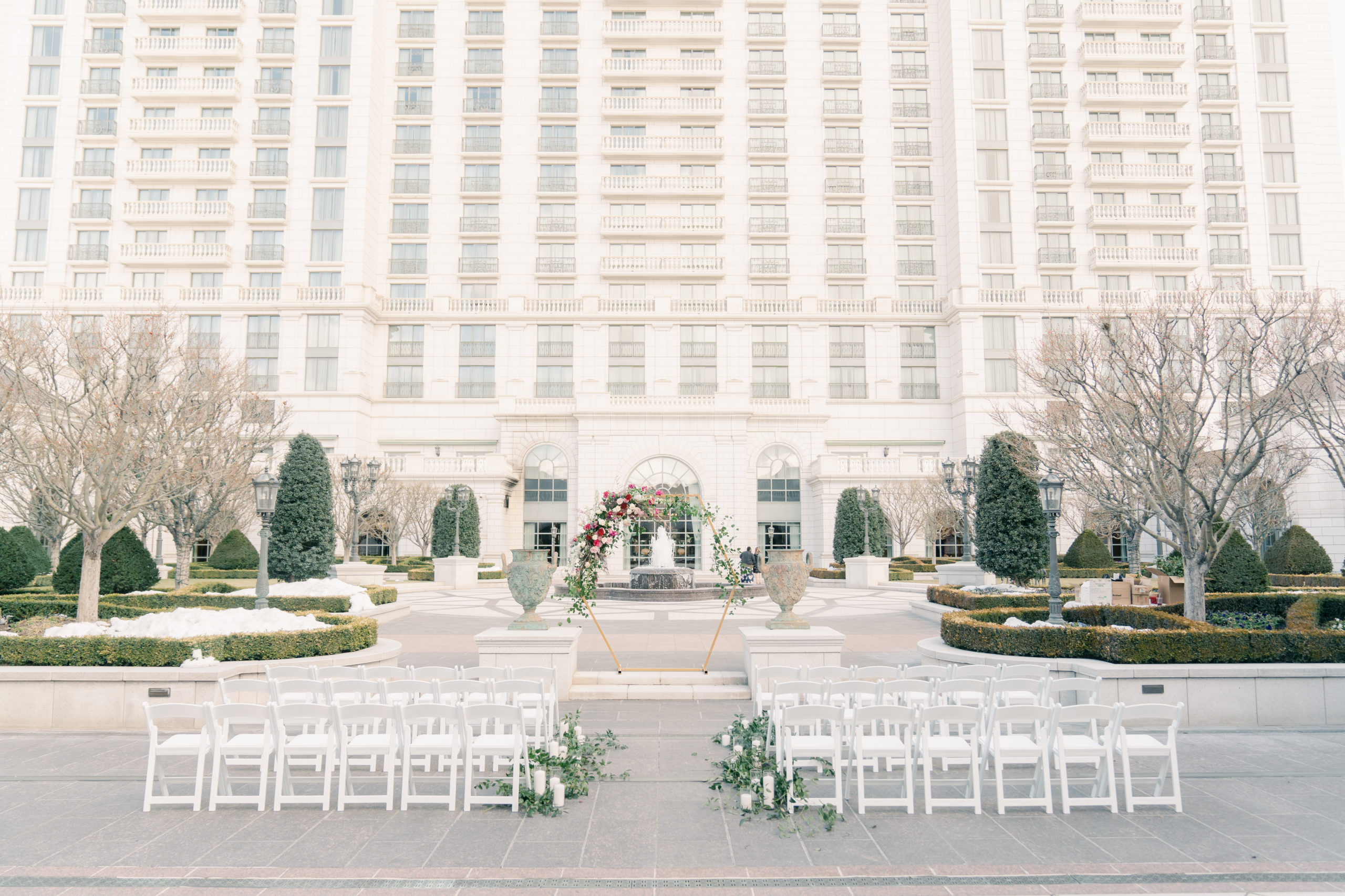 Outdoor wedding ceremony set up with white chairs and greenery in the courtyard.